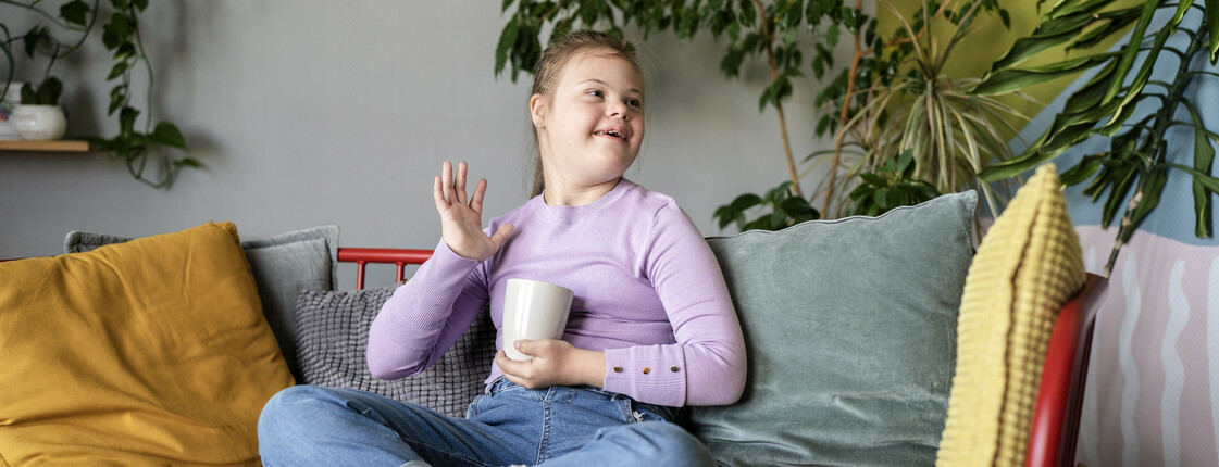 Ein fröhliches Mädchen sitzt entspannt auf einem Sofa, hält eine Tasse und winkt lächelnd.