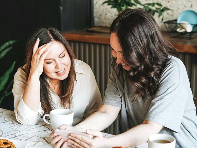 Zwei Frauen sitzen an einem Tisch, trinken Kaffee und schauen gemeinsam auf ein Smartphone, während sie lachen und sich unterhalten.